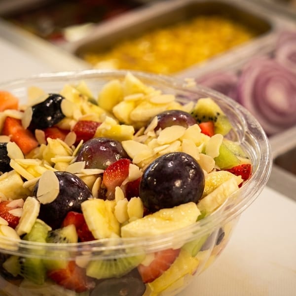 A clear plastic bowl filled with fresh fruit salad, topped with sliced almonds, on a white counter.
