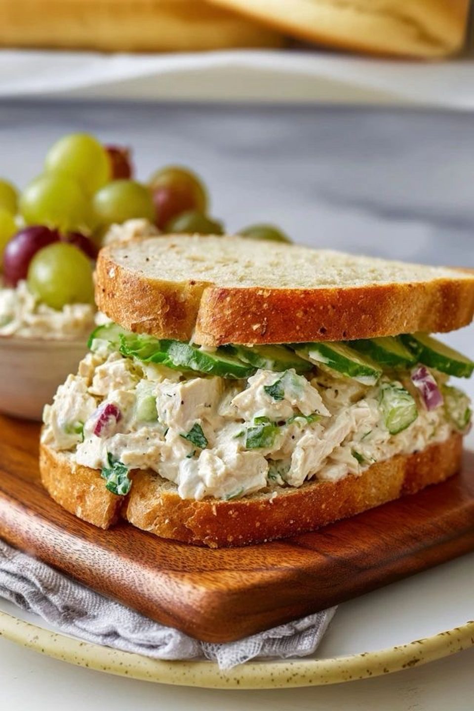 Chicken salad sandwich with cucumber slices on toasted bread, served with grapes on a wooden board.