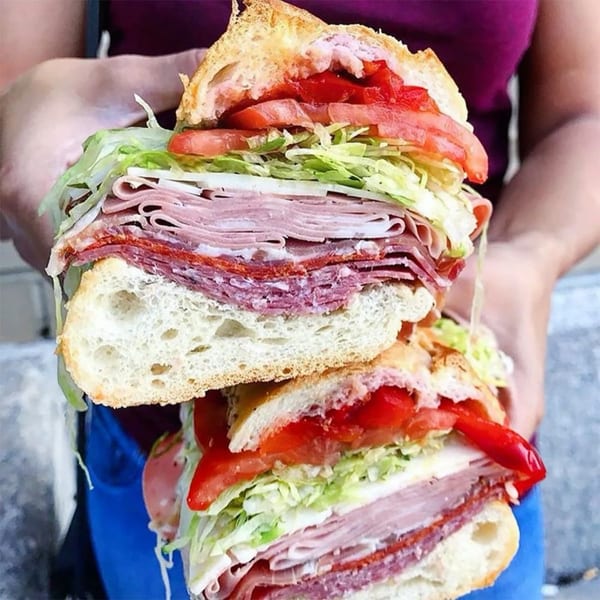 A person holds a large, overflowing deli sandwich filled with meats, cheese, lettuce, and tomatoes.